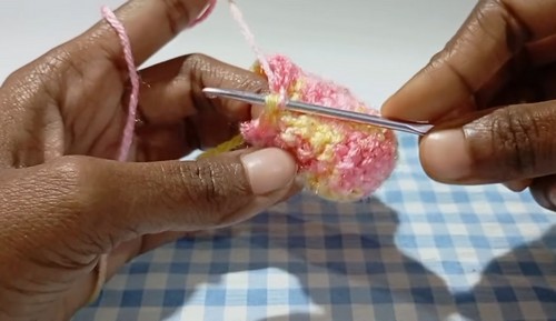Hands using a crochet hook to join two small pastel crocheted heart halves together along the top edge.