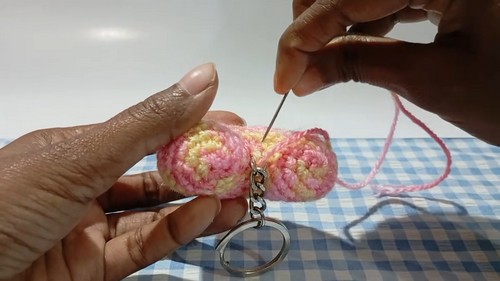 Hands using a tapestry needle to attach a keychain ring to a stuffed pastel multicolor crocheted heart on a blue gingham surface.