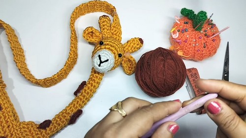 Completed giraffe head and long neck strap laid flat on a white surface next to a ball of dark brown yarn, scissors, and a decorative pumpkin pin cushion.