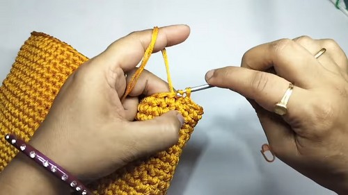Hands using a small crochet hook to work yellow yarn stitches along the top edge of a flat rectangular golden yellow crochet bag panel.