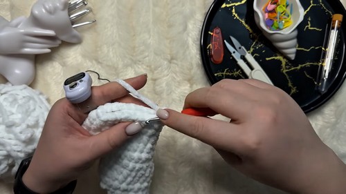 Hands crocheting a white chenille amigurumi wing piece using an orange hook, with craft supplies and a decorative tray visible in the background.