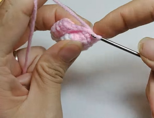 Hands working a crochet hook through a small pink amigurumi piece in progress, with a yarn tail looped overhead.
