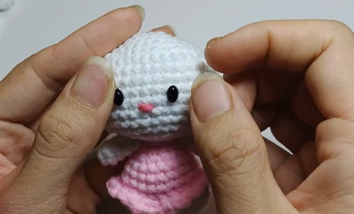 A small white crochet kitten amigurumi with black safety eyes, an embroidered pink nose, and a pink body held between two hands.
