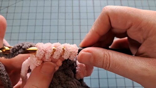 Hands crocheting a pink chenille ear piece directly onto the dark gray mouse head using a gold hook.