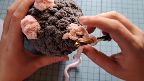 Hands attaching a long pink chenille tail to the back of a dark gray plush crochet mouse body using a gold hook.