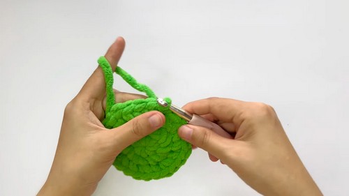 Hands using a white crochet hook to work the opening rounds of a green chenille hat piece, showing the domed shape beginning to form.