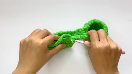 Hands using a white crochet hook to join the two leg sections of a green chenille dungaree piece across the crotch on a white surface.