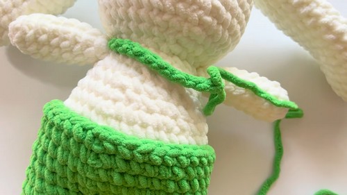 Close-up of a white chenille bunny body mid-construction with green dungaree straps being crocheted across the chest, yarn tail visible at the side.