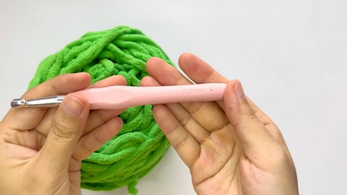 Hands holding a pink crochet hook in front of a large ball of bright green chenille yarn, ready to begin crocheting the frog hat or dungarees.