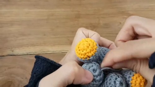 Hands fitting a small round yellow crocheted piece against the gray body of a rhinoceros amigurumi during assembly, with the leg pieces visible against a wooden surface.