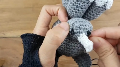 Hands holding a gray crochet rhinoceros amigurumi in progress, with a small white crocheted horn being positioned near the face and black safety eyes already inserted, on a wooden surface
