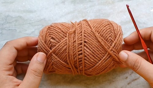 Hands holding a ball of tan brown yarn with a red crochet hook ready to begin the teddy bear keychain pattern