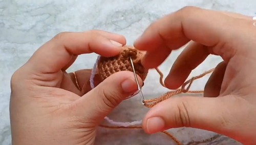 Hands using a tapestry needle to sew a white muzzle piece onto the front of a tiny crocheted brown bear head