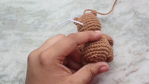Hand holding a small crocheted tan bear body with the head attached and white yarn tails visible showing the assembly stage before adding the muzzle