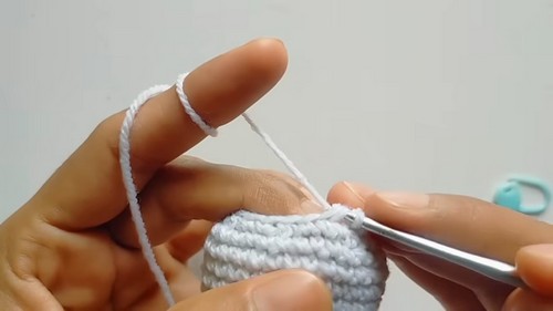 Hands using a silver crochet hook to work light grey and white yarn rounds on a small oval owl body piece with a teal stitch marker visible.