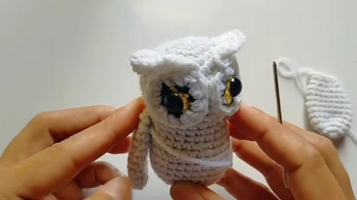 Hands positioning a white crocheted owl amigurumi with yellow and black safety eyes and a fluffy head, with a separate wing piece visible to the right.