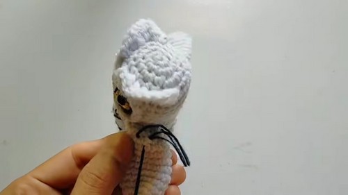 Hand holding a white crocheted owl head from the back, showing the fluffy top feather tuft and black wire talon details below the body.