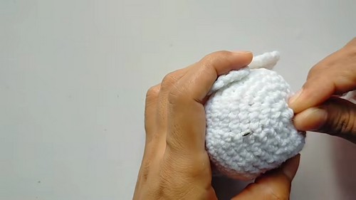 Hands pinning a small crocheted white wing piece onto the side of an owl body using a sewing pin to position it before attaching