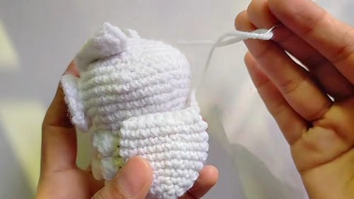 Hands closing the top of a crocheted white owl body with a yarn tail showing the finishing step before adding facial details