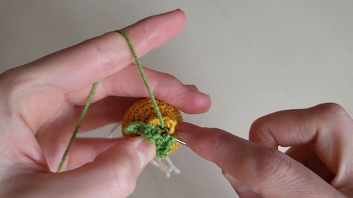 Hands using a tapestry needle to attach a small green crocheted sprout to the top of a golden yellow onion amigurumi, with white root strands visible below.