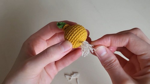 Hands holding a finished golden yellow crocheted onion body with a green sprout on top, threading white yarn root strands through the base.