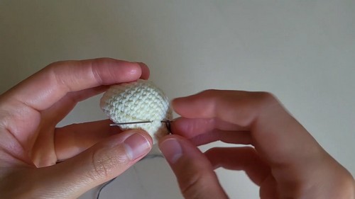Hands inserting a yarn needle through the side of a stuffed cream amigurumi head piece to begin attaching a separate section.