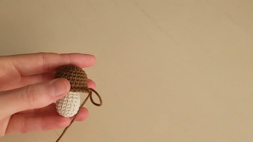 A small two-piece crochet mushroom amigurumi with a brown cap and cream stem held between two fingers, nearly complete with a yarn tail still attached.