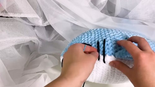 Hands attaching black safety eyes to a light blue and white velvet yarn crochet shark body in progress.