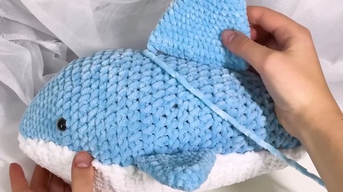 Hands holding a large light blue velvet crochet shark amigurumi with a white belly and attached dorsal fin, partially complete.