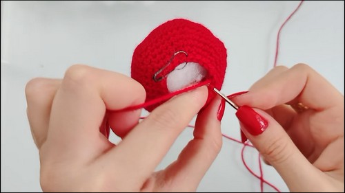 Hands closing the opening of a stuffed red crochet crab body using a tapestry needle and red yarn, with polyfill visible inside.