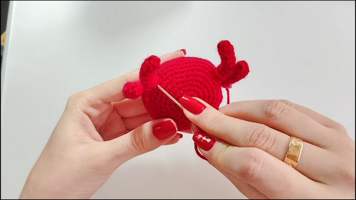 Hands using a tapestry needle to sew a leg onto the underside of a red crocheted crab body, with small claw pieces visible on top.