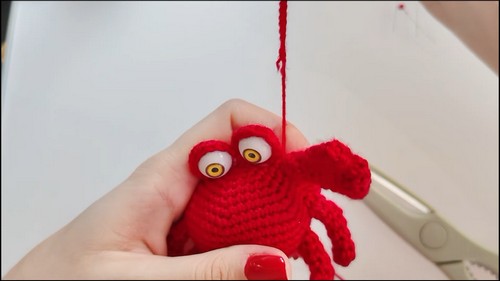 Hand holding a nearly complete red crochet crab amigurumi with yellow safety eyes and attached claws, with a yarn tail extending upward.