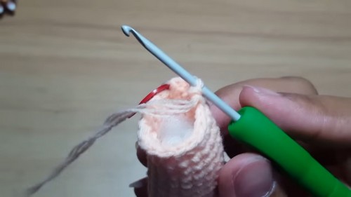 Hands holding a small peach crocheted doll leg section with a red stitch marker and green crochet hook mid-stitch on a wooden table.