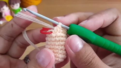 Close-up of hands crocheting peach yarn around a stitch marker on a doll leg piece using a green hook, with the tube shape visible.