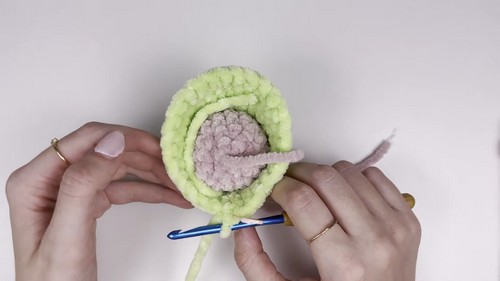 Hands using a blue crochet hook to work light green chenille yarn around the pink inner mouth of a bass amigurumi, showing the open mouth construction in progress.