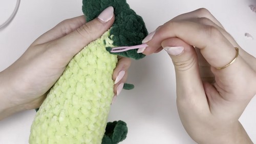 Hands using a pink stitch marker to join a dark green fin piece to the side of a light green chenille bass amigurumi body.