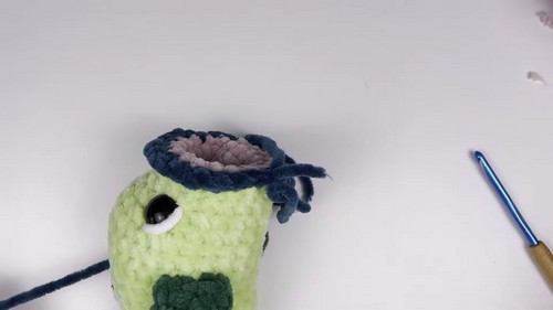 Top-down view of a light green chenille bass amigurumi head with an open teal-rimmed mouth cavity and pink interior, with a blue crochet hook resting nearby.